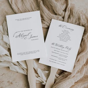 May include: Two white wedding fan programs with wooden handles. The left fan reads "Welcome to the Wedding of Abigail & James" and the date. The right fan lists the ceremony order and wedding party names.