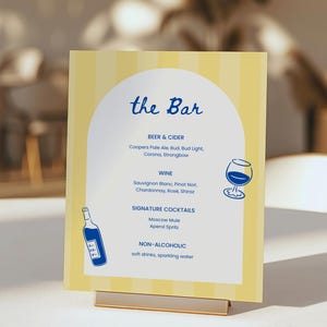 May include: A yellow and white striped bar menu with the text "the Bar" in blue script. The menu lists beer, cider, wine, signature cocktails, and non-alcoholic beverages. A gold metal stand holds the menu.