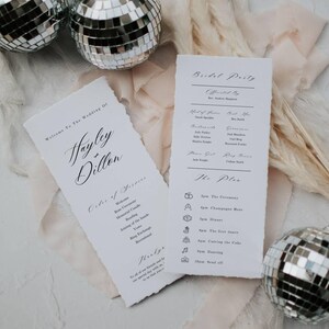 Pode incluir: Ordem do serviço da cerimônia de casamento e informações sobre a festa de casamento impressas em dois cartões brancos com borda rasgada. Os cartões estão sobre uma superfície rosa claro com duas bolas de discoteca prateadas.