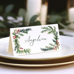 Puede incluir: Una tarjeta de lugar blanca con un diseño de acuarela verde y rojo de ramas de pino y bayas. El nombre "Angelina" está escrito en cursiva negra.