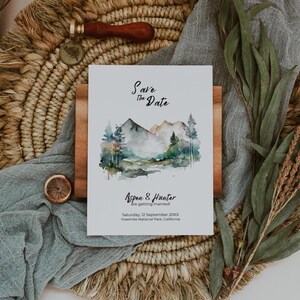 May include: Save the date card featuring a watercolour illustration of a mountain range with a river flowing through it. The text reads "Save the Date" and "Aspen & Hunter are getting married!" with the date and location below.