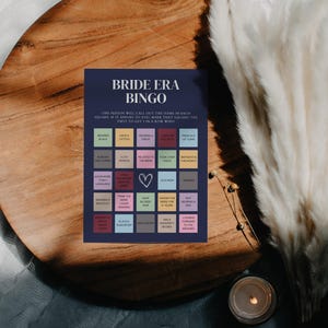 K&ouml;nnte beinhalten: Eine blaue und wei&szlig;e Bingokarte mit dem Titel "Bride Era Bingo". Die Karte hat 25 Felder mit verschiedenen S&auml;tzen, die mit einer Hochzeit zu tun haben, wie z. B. "Schwarz tragen", "ein Tattoo haben", "ein Kleid tragen", "gibt das Alter von", "ausserhalb der Stadt", "bereits ein Baby gehabt", "eine Katzenperson", "mit der Braut verwandt", "heute ein Nickerchen gemacht", "Geburtstag ist der Monat", "mehr als eine Sprache sprechen", "ein Armband tragen", "denke, dass die Braut toll aussieht", "blonde Haare haben", "die Braut seit &uuml;ber 5 Jahren kennen", "keinen Ring tragen", "die Braut heute kennengelernt", "einen Mannschaftssport spielen", "einen Master haben", "einen Hochschulabschluss haben", "sich auf die Hochzeit freuen".