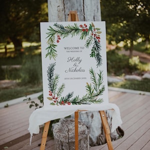 Puede incluir: Un cartel de bienvenida de boda blanco con una ilustración de acuarela de ramas de pino verdes y bayas rojas. El cartel dice "Bienvenido a la boda de Holly & Nicholas 18 de diciembre de 20XX".