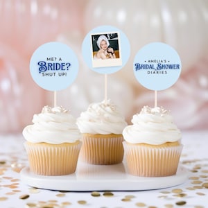 以下が含まれることがあります： 白いフロスティングと金色のスプリンクルをトッピングした3つのカップケーキ。青い円形のカップケーキトッパーが飾られています。トッパーには「ME? A BRIDE? SHUT UP!」と「AMELIA'S BRIDAL SHOWER DIARIES」の文字と、女性の画像が描かれています。