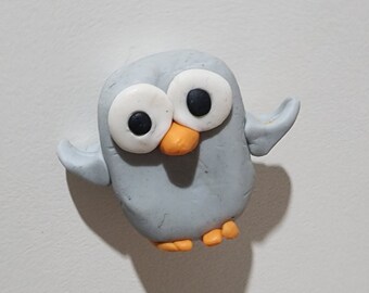 May include: A grey owl figurine with large white eyes and an orange beak. The owl is made of clay and has a whimsical expression.