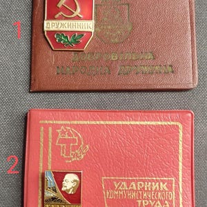 Può includere: Due spille vintage dell'era sovietica. La spilla superiore è marrone con un emblema rosso con falce e martello e la scritta "ДРУЖИННИК". La spilla inferiore è rossa con un emblema dorato di Lenin e la scritta "УДАРНИК КОММУНИСТИЧЕСКОГО ТРУДА".