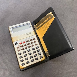 May include: A vintage gold and beige calculator, model MK 51, with a black leather case. The calculator has numerous buttons and a digital display. A yellow and white booklet is visible inside the case. The text "Электроника МК 51" is printed on the calculator.