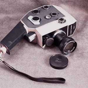 May include: Vintage Super 8 film camera with a black and white body, black lens, and a black hand strap. The camera has several dials and buttons on top and a lens cap. The camera is on a gray surface.