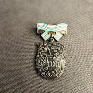 Op de afbeelding: Een zilverkleurige metalen medaille met een lichtblauw en wit gestreepte strik aan de bovenzijde. De medaille toont een moeder met een kind, met de cyrillische tekst "МАТЕРИНСКАЯ СЛАВА III" en "СССР" naast een hamer en sikkel symbool.