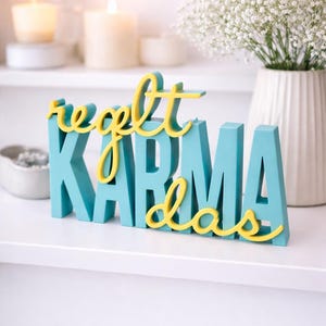 Könnte beinhalten: Ein türkisfarbenes und gelbes Holzschild mit der Aufschrift "regelt karma ideas". Die Buchstaben sind türkis, mit den Wörtern "regelt" und "ideas" in gelber Schrift. Das Schild steht auf einem weißen Regal.