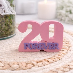 Könnte beinhalten: Eine rosa Holzfigur mit der Zahl 29 und dem lila Wort „FOREVER“. Das Dekorationselement steht auf einem gewebten Platzset. Kerzen und ein Glas mit Grünzeug sind im Hintergrund.