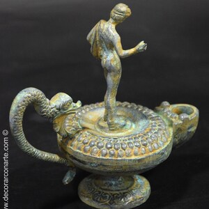May include: A bronze oil lamp with a green patina. The lamp features a figure of a woman standing on a circular base. The lamp has a long, curved handle shaped like a serpent's head.