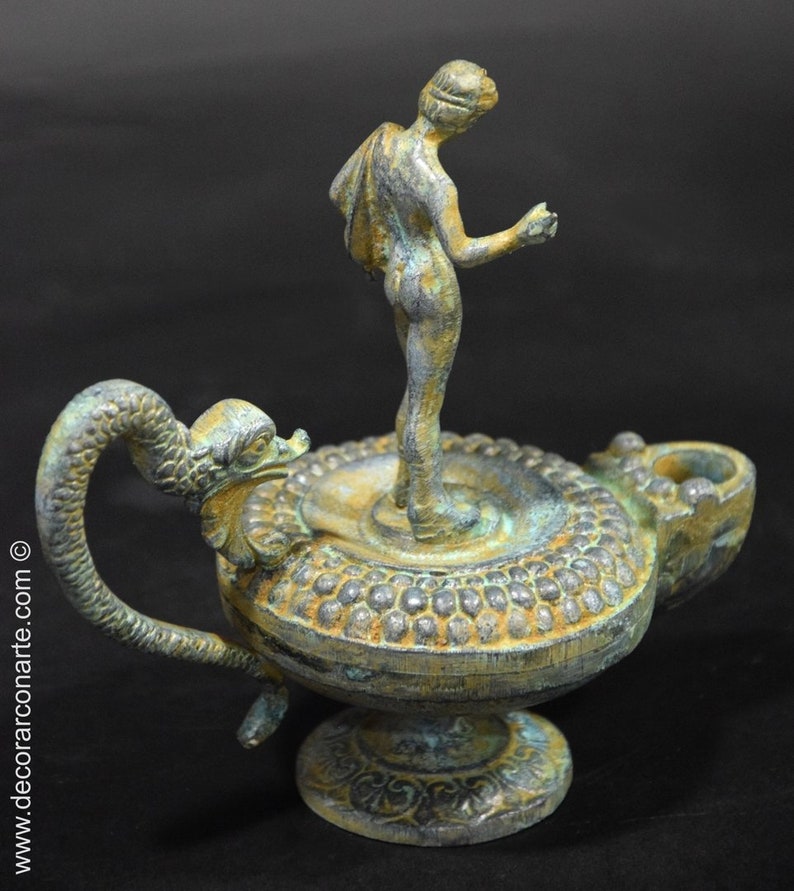 May include: A bronze oil lamp with a green patina. The lamp features a figure of a woman standing on a circular base. The lamp has a long, curved handle shaped like a serpent's head.