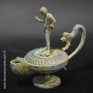 May include: A bronze oil lamp with a decorative dragon handle and a small standing figure on top. The lamp has a green patina and intricate details.