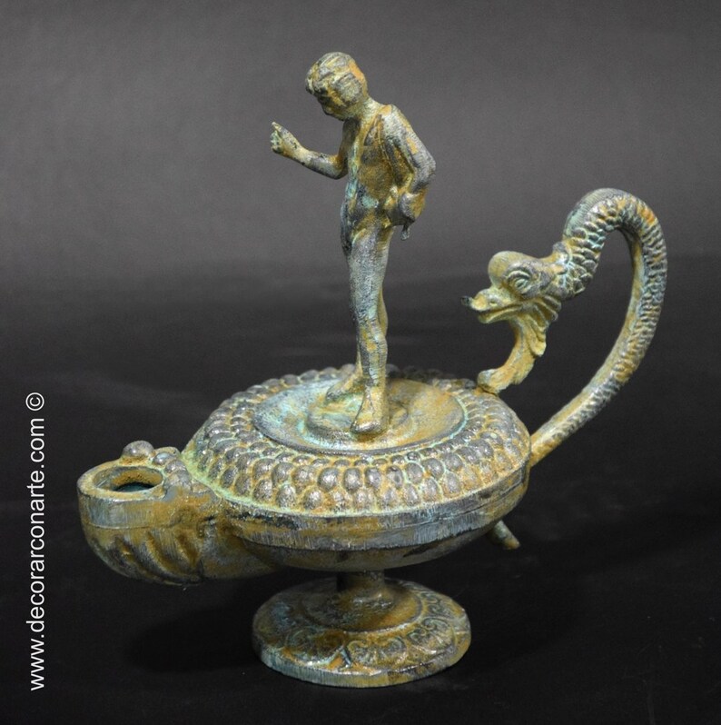 May include: A bronze oil lamp with a decorative dragon handle and a small standing figure on top. The lamp has a green patina and intricate details.