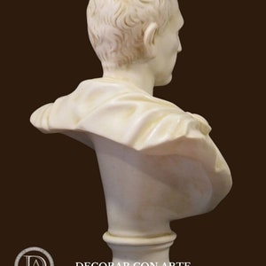 May include: A cream-coloured marble bust of a person, seen from the side and back. The sculpture has detailed hair and draped clothing. The bust is mounted on a round, fluted pedestal. The background is a solid brown.