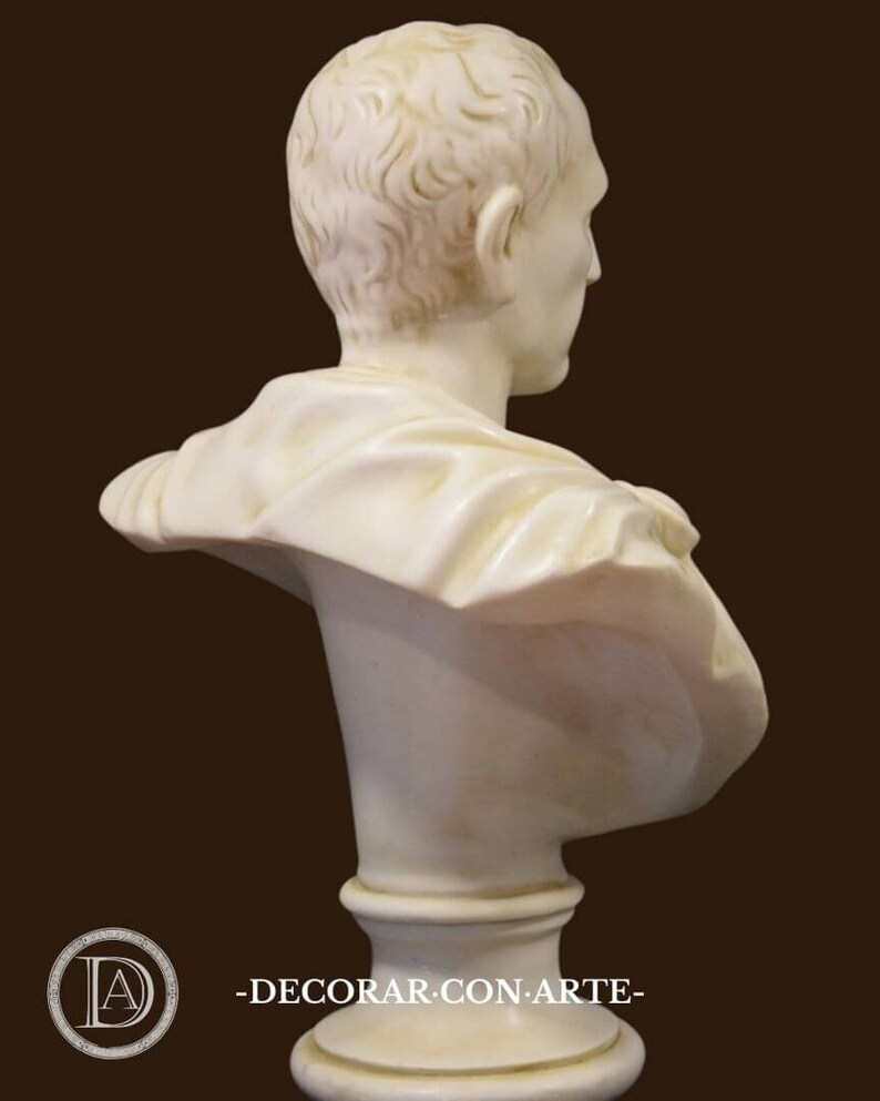May include: A cream-coloured marble bust of a person, seen from the side and back. The sculpture has detailed hair and draped clothing. The bust is mounted on a round, fluted pedestal. The background is a solid brown.