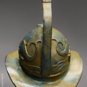 May include: A bronze helmet with a decorative design and a tall, curved crest. The helmet is a dark green and gold color.