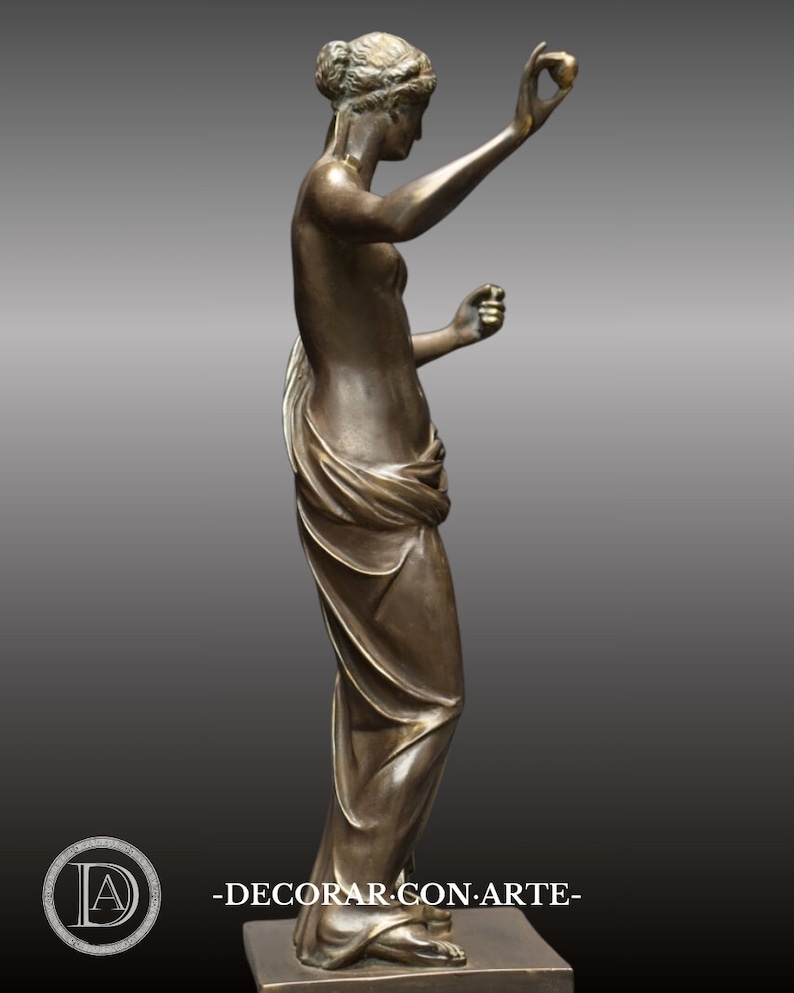 May include: A bronze sculpture of a woman standing with her arms raised, holding a small object in each hand. The woman is wearing a flowing robe and has a serene expression on her face.