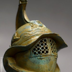 May include: A bronze gladiator helmet with a decorative crest and a face mask with a grid pattern. The helmet has a green patina and is on a display stand.