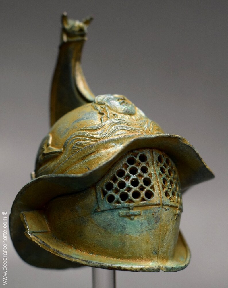 May include: A bronze gladiator helmet with a decorative crest and a face mask with a grid pattern. The helmet has a green patina and is on a display stand.