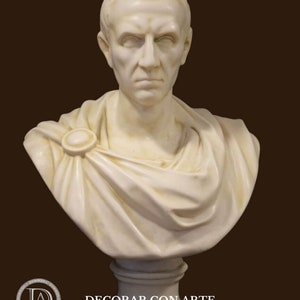 May include: A cream-coloured marble bust of a man with short hair and a stern expression. The bust features a draped garment secured with a circular clasp. The base is a simple, cylindrical pedestal. The text "-DECORAR.CON.ARTE-" is at the bottom.
