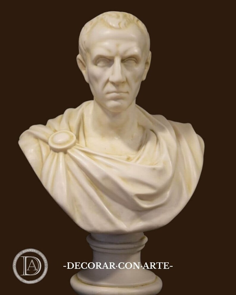 May include: A cream-coloured marble bust of a man with short hair and a stern expression. The bust features a draped garment secured with a circular clasp. The base is a simple, cylindrical pedestal. The text "-DECORAR.CON.ARTE-" is at the bottom.
