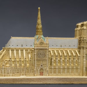 May include: A detailed miniature replica of Notre Dame Cathedral in Paris, France. The model is made of a light brown material and features intricate details, including the iconic spire and stained glass windows.