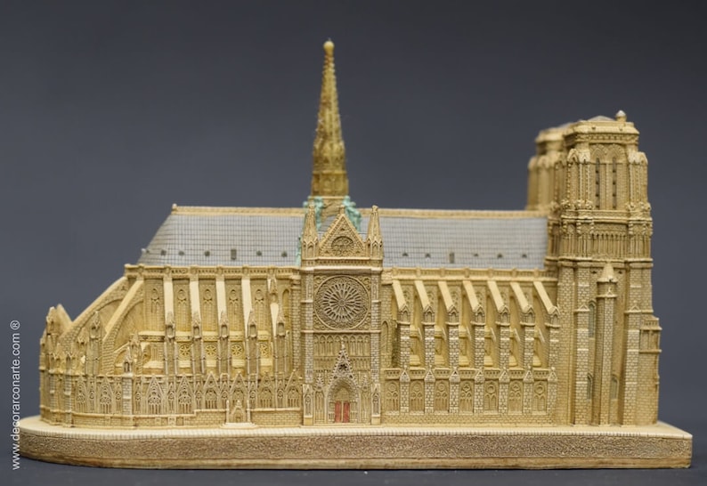 May include: A detailed miniature replica of Notre Dame Cathedral in Paris, France. The model is made of a light brown material and features intricate details, including the iconic spire and stained glass windows.