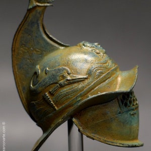 May include: A bronze helmet with a dolphin design and a face mask. The helmet has a tall, pointed crest and a decorative design on the side.