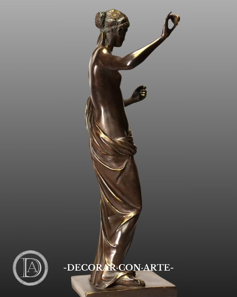 May include: Bronze sculpture of a woman standing on a base. She is wearing a flowing robe and holding a small object in her right hand. The sculpture is in a classic style.