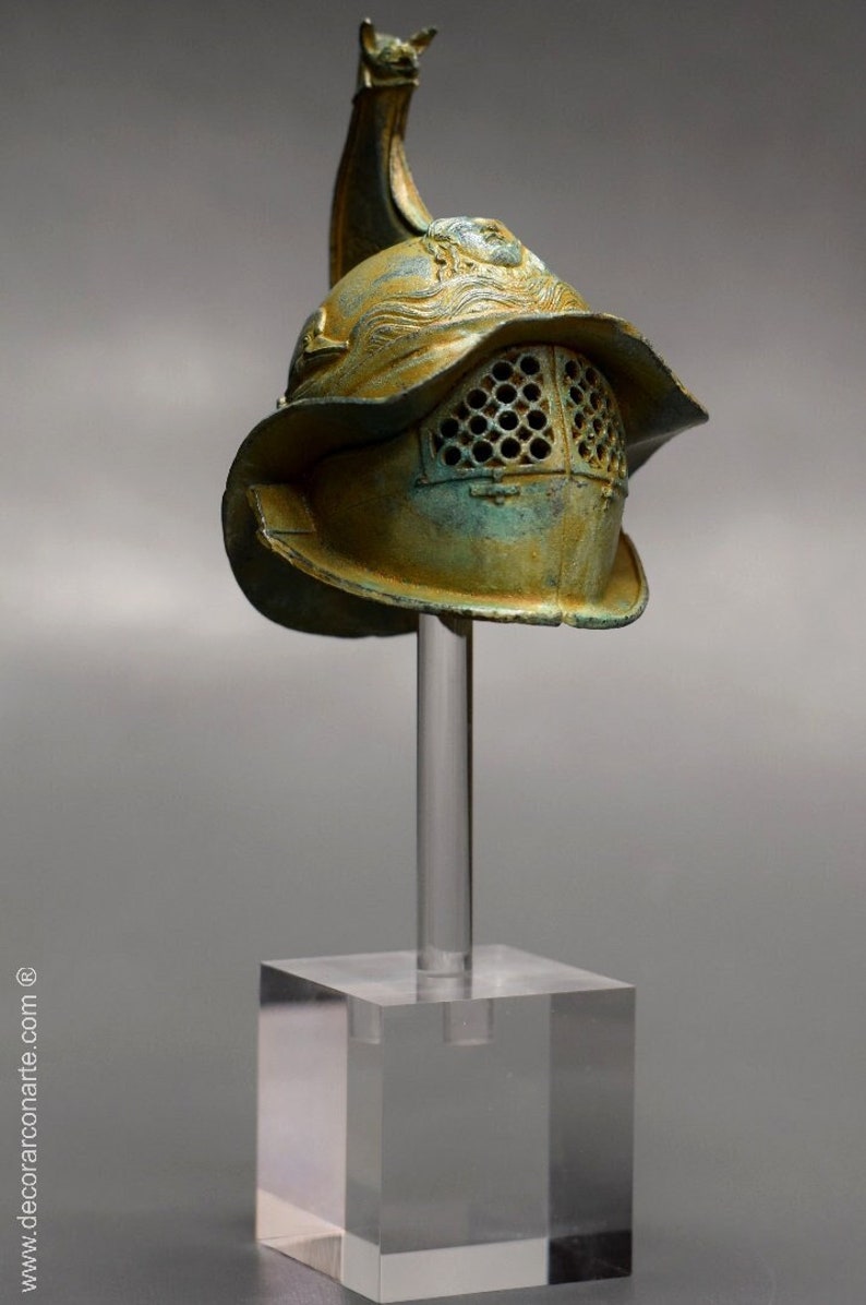 May include: A bronze gladiator helmet with a decorative crest and a wire mesh face guard. The helmet is displayed on a clear acrylic stand.