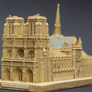May include: A detailed miniature replica of the Notre Dame Cathedral in Paris, France. The model is made of a light brown material and features intricate details, including the iconic twin towers, gargoyles, and stained glass windows. The base of the model is inscribed with the words "NOTRE-DAME DE PARIS."