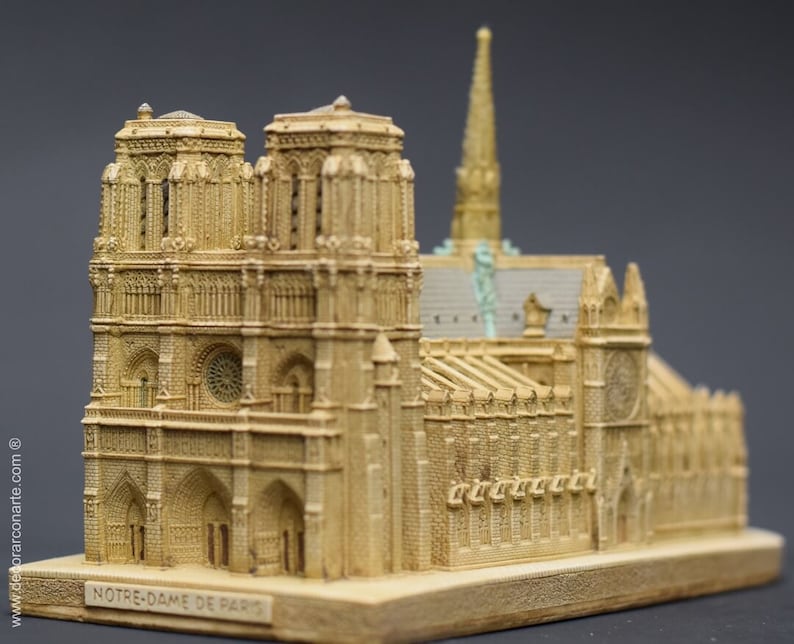 May include: A detailed miniature replica of the Notre Dame Cathedral in Paris, France. The model is made of a light brown material and features intricate details, including the iconic twin towers, gargoyles, and stained glass windows. The base of the model is inscribed with the words "NOTRE-DAME DE PARIS."