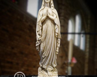 May include: A cream-colored statue of a woman with clasped hands, wearing a draped robe and veil. A rosary hangs from her hands. The statue is on a marble base, with the text "-DECORAR CON ARTE-" at the bottom.