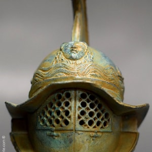 May include: A bronze Roman gladiator helmet with a face sculpted on the top and a wire mesh face guard. The helmet is a dark green and gold color.