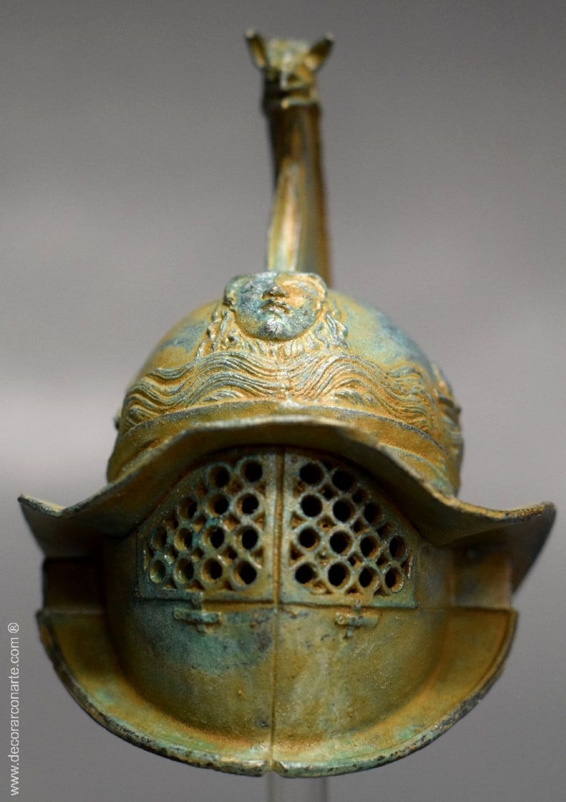 May include: A bronze Roman gladiator helmet with a face sculpted on the top and a wire mesh face guard. The helmet is a dark green and gold color.