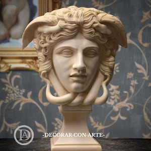 May include: A cream-colored Medusa head sculpture on a pedestal. The sculpture features detailed facial features, flowing hair, and serpent-like elements. The background includes a framed painting and a patterned wall. The text "-DECORAR CON ARTE-" is visible.
