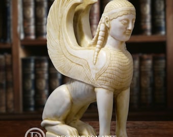 May include: Ivory-coloured sphinx sculpture with detailed wings and a serene expression. The sculpture features intricate carvings and is set against a backdrop of a bookshelf. The base of the sculpture has the text "-DECORAR CON ARTE-".