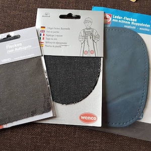Könnte beinhalten: Zwei Packungen Bügel-Flicken für Kleidung. Eine Packung enthält zwei schwarze Stoff-Flicken und die andere einen blauen Leder-Flicken. Der Text auf der Packung lautet "Flecken zum Aufbügeln" und "Leder-Flecken aus echtem Nappaleder vorgelocht waschbar 30°".