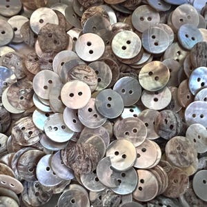 Könnte beinhalten: Eine große Sammlung runder Zweilochknöpfe aus natürlichem Muschelmaterial. Die Knöpfe weisen verschiedene irisierende, graue und braune Töne mit einzigartigen Wirbelmustern und glatten Oberflächen auf und sind in einem dichten Haufen angeordnet.
