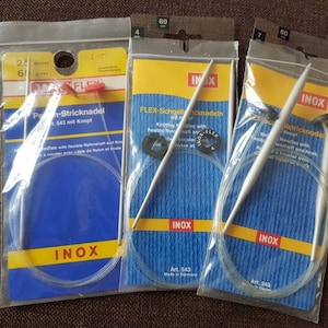Könnte beinhalten: Drei Packungen INOX Stricknadeln. Jede Packung hat ein blau-gelbes Etikett mit Text und ein transparentes Kunststofffenster, das die Nadeln zeigt. Die Nadeln sind silberfarben mit weißen Spitzen und durch ein klares, flexibles Kabel verbunden.