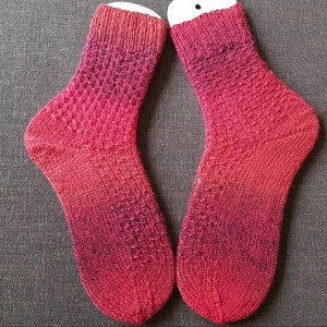 Könnte beinhalten: Ein Paar handgestrickte Socken in einem rot-schwarzen Farbverlauf. Die Socken haben ein strukturiertes Strickmuster und liegen flach auf einer grauen Stoffoberfläche.