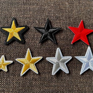 Könnte beinhalten: Eine Sammlung von sternförmigen Aufnähern in verschiedenen Farben, darunter Gelb, Schwarz und Rot. Die Aufnäher haben gestickte Details und sind auf einer braunen, strukturierten Oberfläche angeordnet. Einige Sterne haben einen schwarzen Rand.