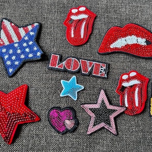 Könnte beinhalten: Eine Sammlung von Pailletten-Patches in verschiedenen Formen und Designs. Enthält einen Stern mit amerikanischem Flaggenmuster, rote Lippen, einen "LOVE"-Patch, Sterne, ein Herz und einen Lippenstift.