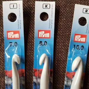 Könnte beinhalten: Drei weiße Häkelnadeln mit blauer und weißer Verpackung. Die Häkelnadeln sind mit der Größe und der Marke "Prym" gekennzeichnet.