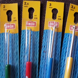 May include: Four different sized crochet hooks in individual packaging. The hooks are labeled with their size in millimeters: 2 mm, 2.5 mm, 3 mm, and 3.5 mm. The packaging is yellow with blue and white yarn inside. The hooks are made of metal and have a silver color. The packaging also includes the brand name "INOX" and the article number "235".