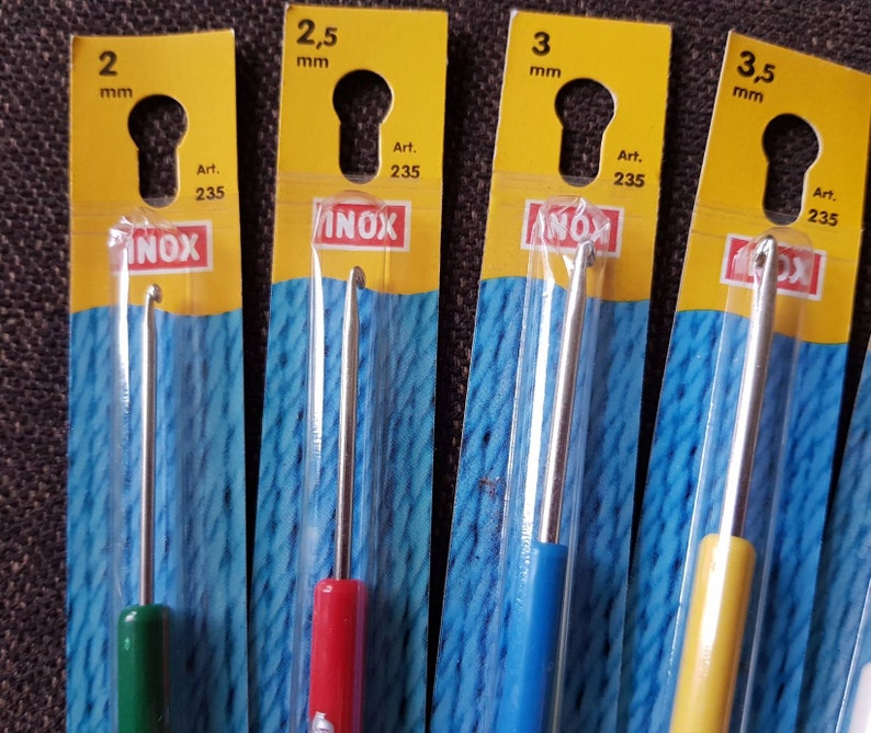 May include: Four different sized crochet hooks in individual packaging. The hooks are labeled with their size in millimeters: 2 mm, 2.5 mm, 3 mm, and 3.5 mm. The packaging is yellow with blue and white yarn inside. The hooks are made of metal and have a silver color. The packaging also includes the brand name "INOX" and the article number "235".