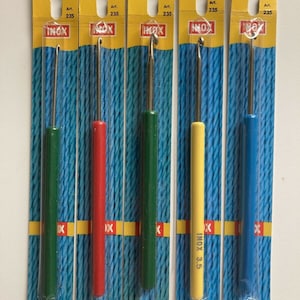 May include: Five different sized crochet hooks in individual plastic packaging. The hooks are labeled with their size in millimeters: 2 mm, 2.5 mm, 3 mm, 3.5 mm, and 4.5 mm. The hooks are all made of metal and have a silver finish. The packaging is yellow with blue and red accents. The brand name "INOX" is printed on the packaging.