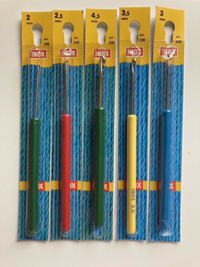 May include: Five different sized crochet hooks in individual plastic packaging. The hooks are labeled with their size in millimeters: 2 mm, 2.5 mm, 3 mm, 3.5 mm, and 4.5 mm. The hooks are all made of metal and have a silver finish. The packaging is yellow with blue and red accents. The brand name "INOX" is printed on the packaging.
